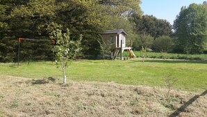 Een klein gebouw met een ladder en een glijbaan, omringd door een grasveld en bomen.