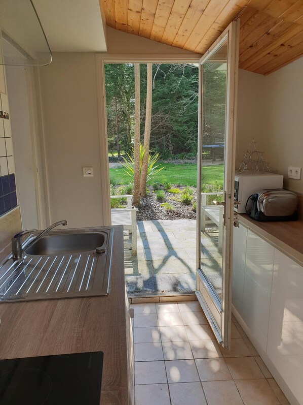 A kitchen with a sink, a toaster, and a view of a garden through an open door. - Warm house in the heart of a forest, close to shopping and beaches (Fouesnant)