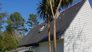 A small white building with a sloped roof, a bench outside, and a garden area with various plants. - Warm house in the heart of a forest, close to shopping and beaches (Fouesnant)