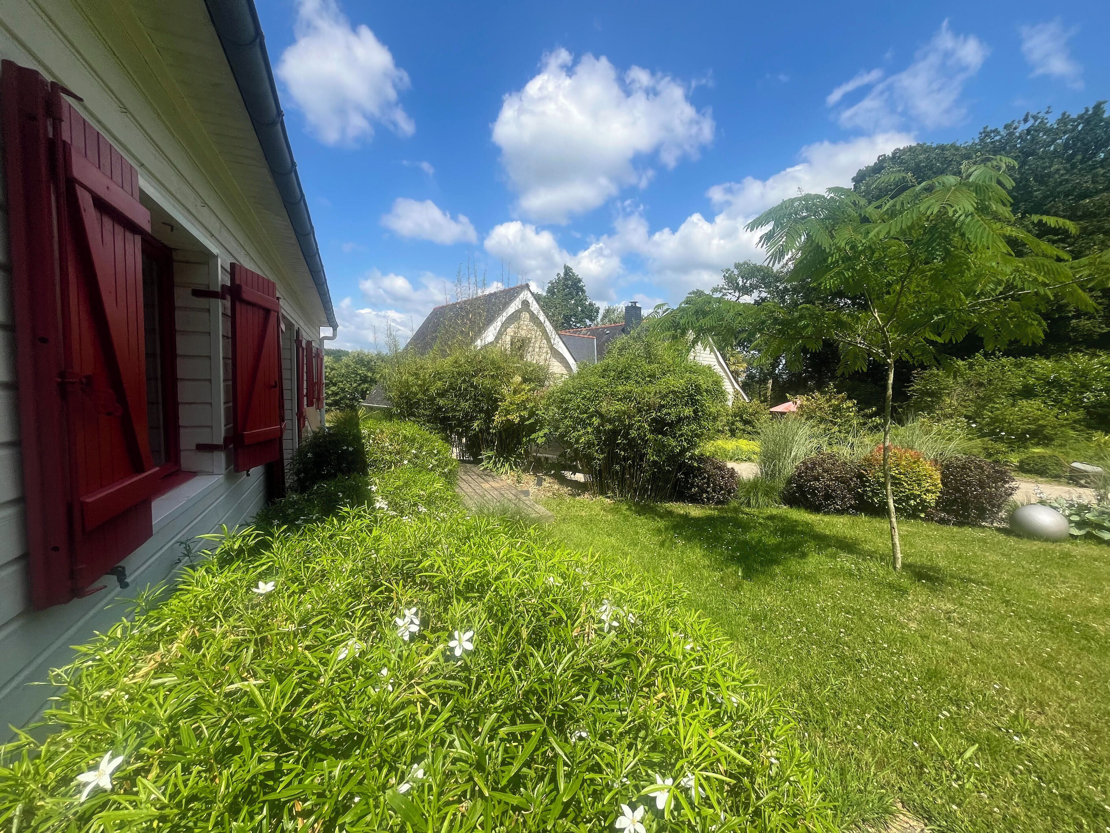 Un jardín con una casa, contraventanas rojas y un cielo azul despejado.