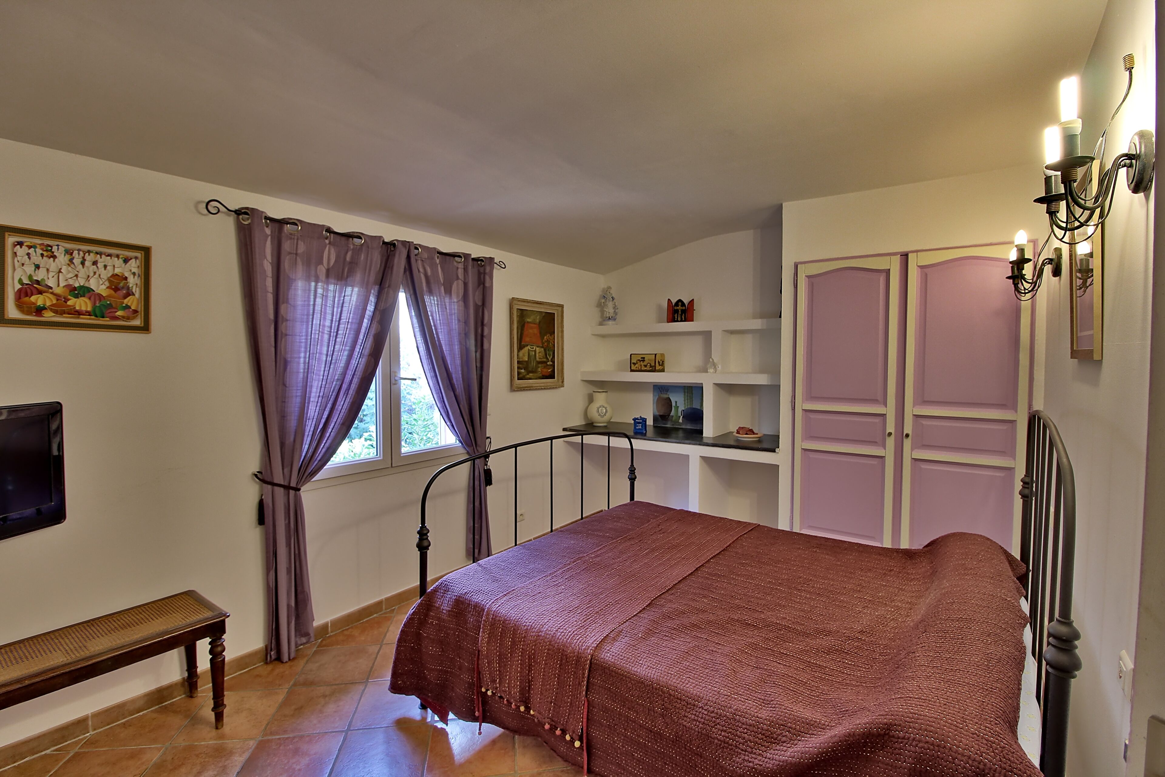A bedroom with a bed, a window with curtains, a bedside table, a TV, and a shelf with decorative items.
