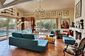 Ein großzügiges Wohnzimmer mit einer breiten Couch, einem Sessel und einem Kamin. An den Wänden befinden sich mehrere gerahmte Bilder und von der Decke hängt ein Chandelier.