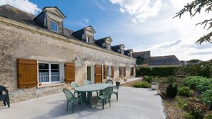 A building with a stone facade, multiple windows with wooden shutters, and a paved patio with a round table and chairs.