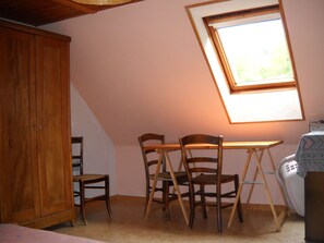 Een kamer met een schuine balkenlaag, een houten tafel met stoelen, een kledingkast en een raam met uitzicht op groen.