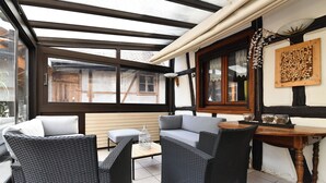 A covered patio area with wicker furniture, a wooden table, and a view of the street. - Manala ***, Alsatian house for 7 to 9 people in Kaysersberg - 3 bedrooms, 2 bathrooms, veranda (Kaysersberg-Vignoble)