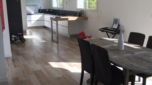 Une cuisine moderne avec des meubles blancs, un coin repas doté d’une table et de chaises en bois, et une vue sur l’extérieur grâce à de grandes fenêtres.
