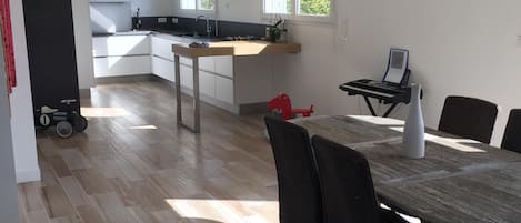A modern kitchen with white cabinets, a dining area with a wooden table and chairs, and a view of the outdoors through large windows.
