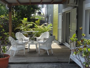 Een overdekt terras met houten vlonder, wit rieten meubilair en potplanten.