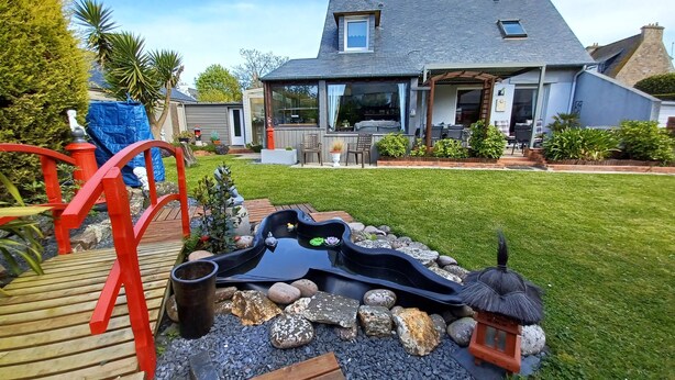 A house with a backyard containing a wooden deck, a small pond with rocks, and a thatched structure.