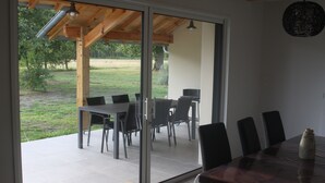 Un comedor con una gran mesa y sillas, techo de madera y vista a un espacio exterior con césped a través de puertas de vidrio.