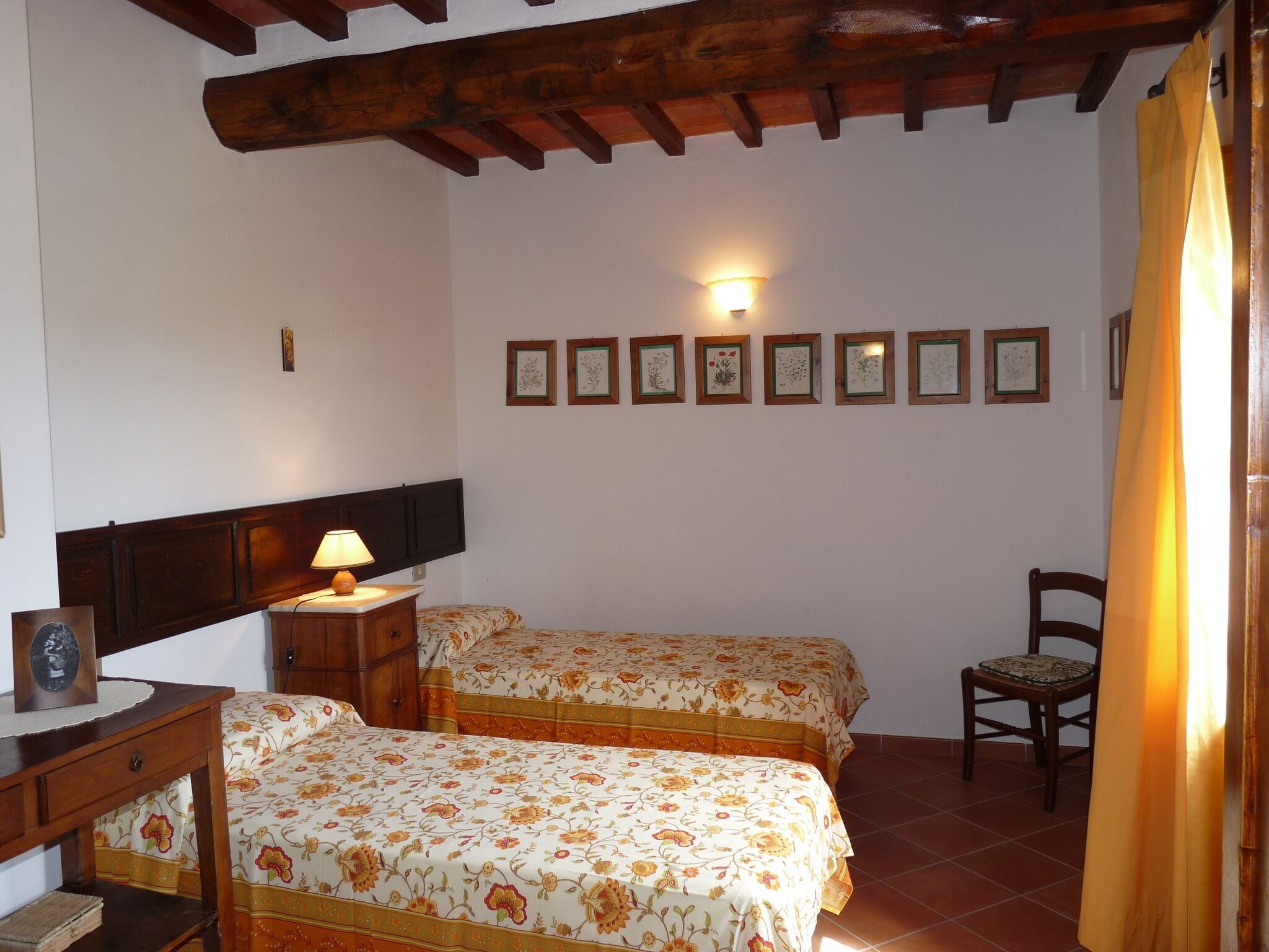 A bedroom with two beds, a wooden headboard, a bedside table with a lamp, a chair, and a wall with framed pictures.