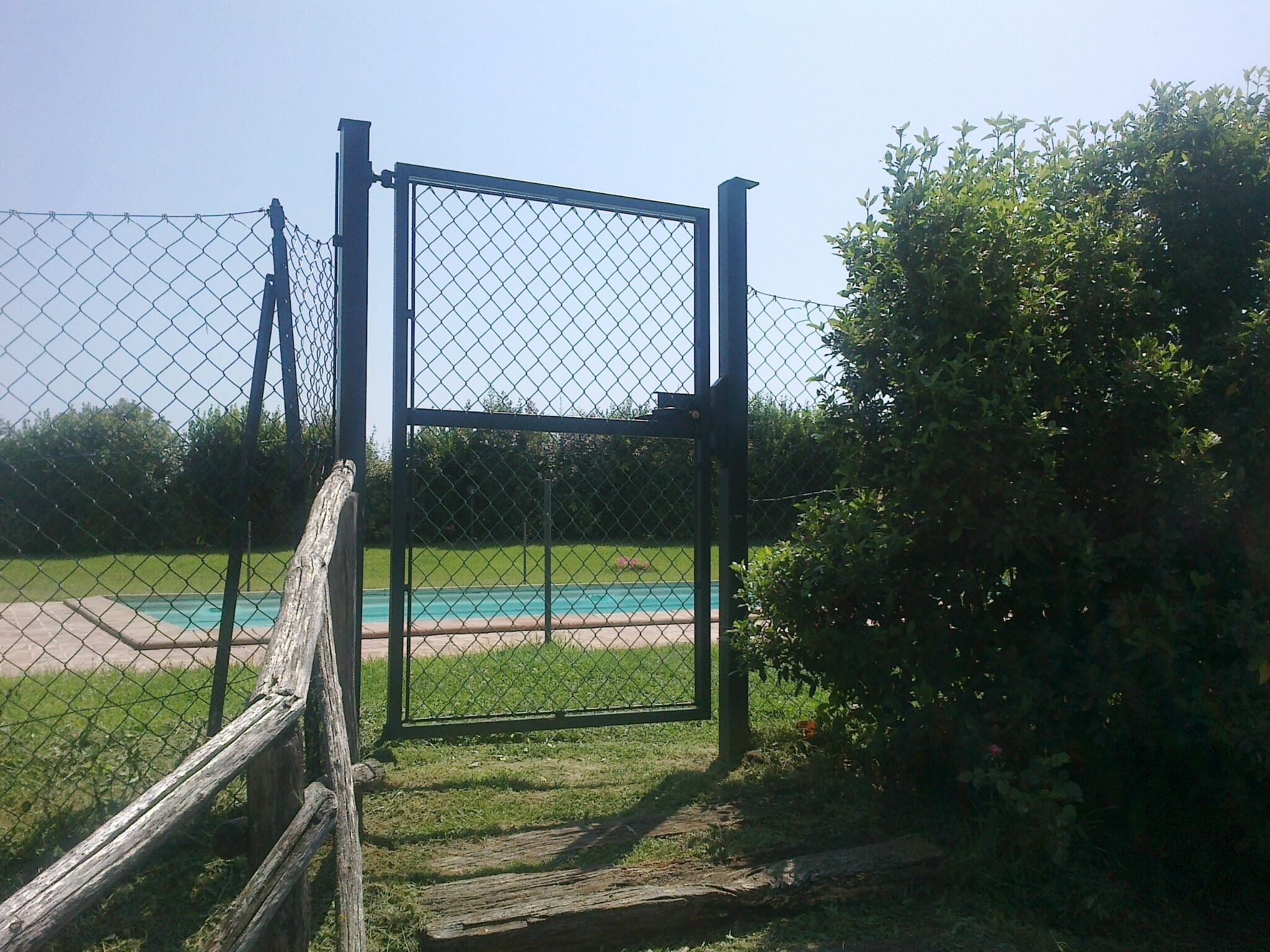 Ein Maschendrahtzaun mit einem Metalltor, ein Holzzaun zur Linken und ein Schwimmbecken im Hintergrund.
