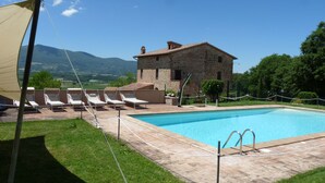 Una piscina con sillones de descanso, un edificio de piedra y vistas a montañas.