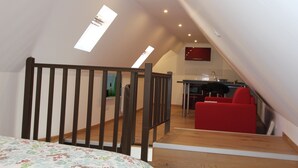 Une chambre à coucher aménagée en mezzanine, avec un lit en bois, une chaise rouge et un coin cuisine à l’arrière-plan.