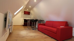 Un espace de vie moderne avec un canapé rouge, un plancher en bois, un coin cuisine et un plafond doté d’un éclairage encastré.