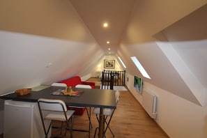 Een compacte, goed verlichte kamer met een eettafel, stoelen, een rode bank en een kleine kitchenette.