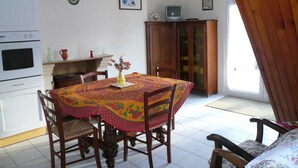 A dining area with a table covered by a colorful tablecloth, surrounded by wooden chairs. A small television is placed on a cabinet above the table. - Small corner for sinners and hikers, calm relaxation in the country. (Saint-Épain)