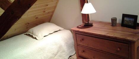 A small, cozy bedroom with a sloped ceiling, a wooden dresser, a bedside lamp, and a single bed with a pillow.
