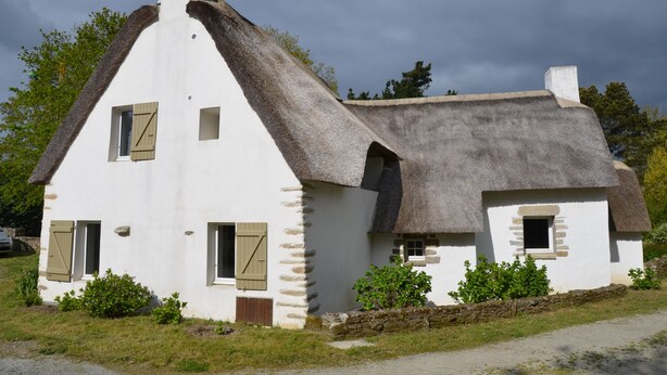 Una casa tradicional de color blanco con techo de paja, muros de piedra y contraventanas de madera.