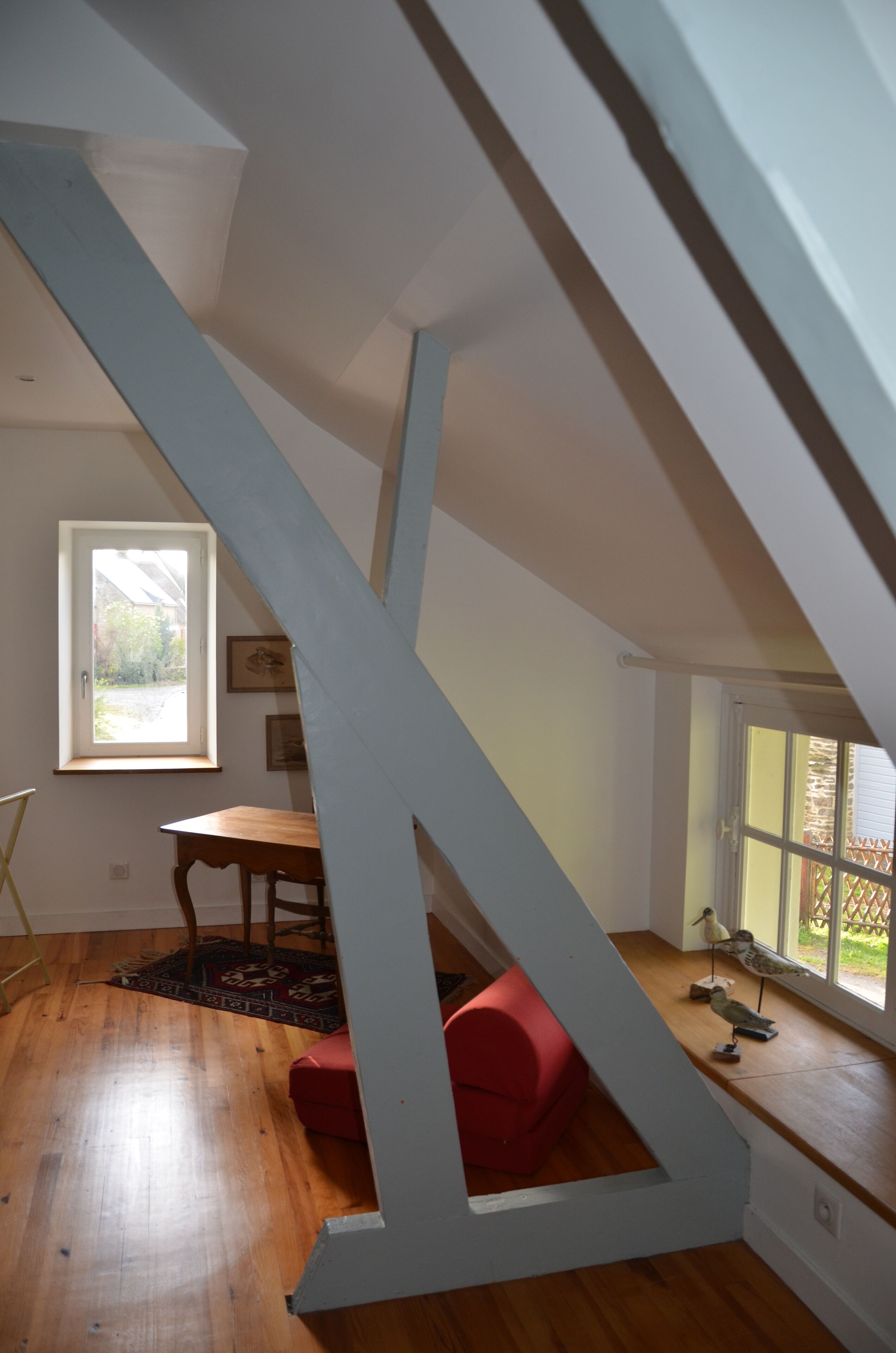 A room with a wooden floor, a red chair, a wooden table, a window with a view of greenery, and a staircase with white railings.