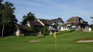 En golfbane med et flag i forgrunden, omgivet af velplejet græs og sandbunkere. I baggrunden ses to store bygninger i traditionel stil med mange vinduer og en balkon.