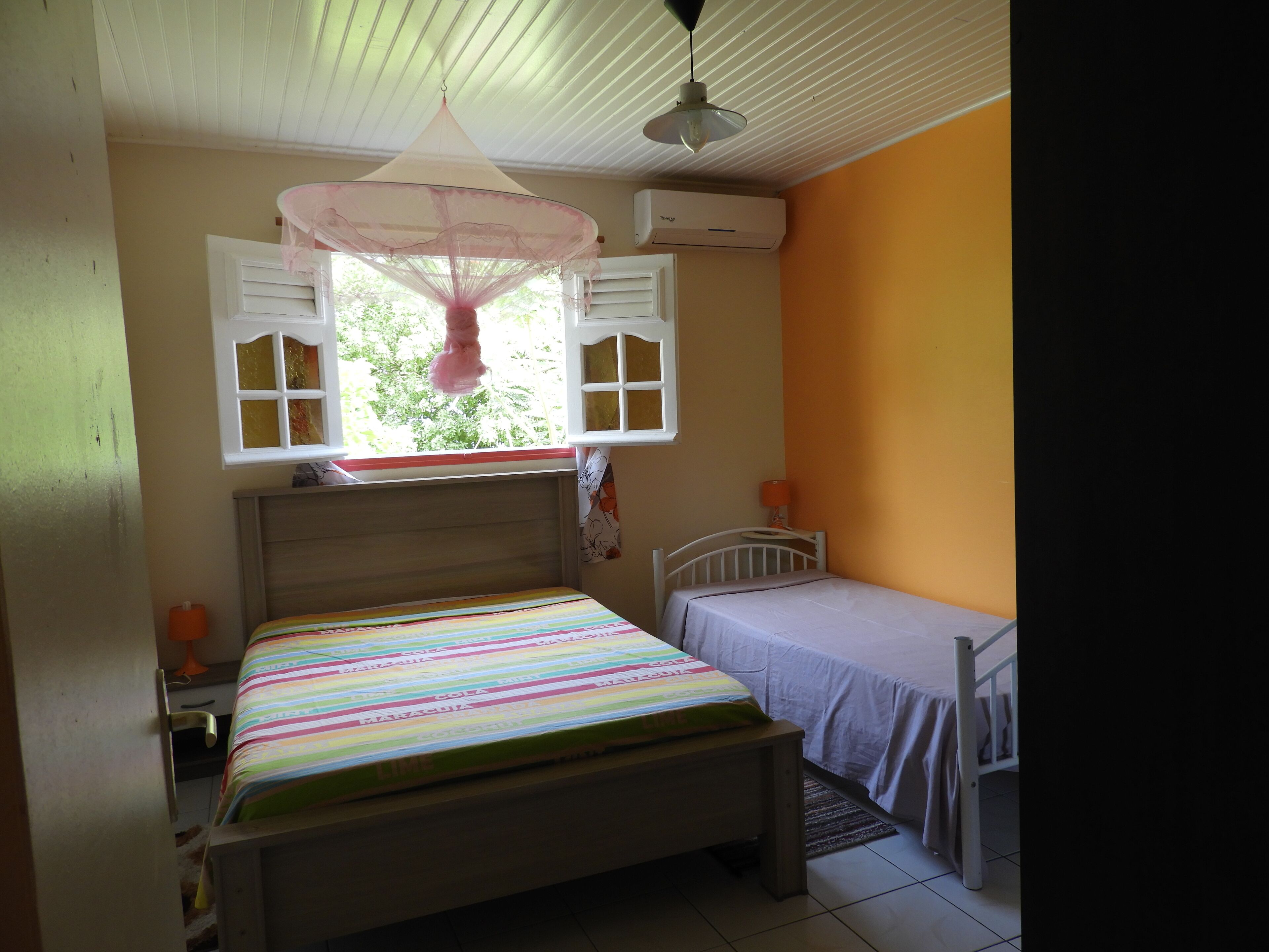 A bedroom with a bed, a bedside table, a window with shutters, an air conditioning unit, and a hanging mosquito net.