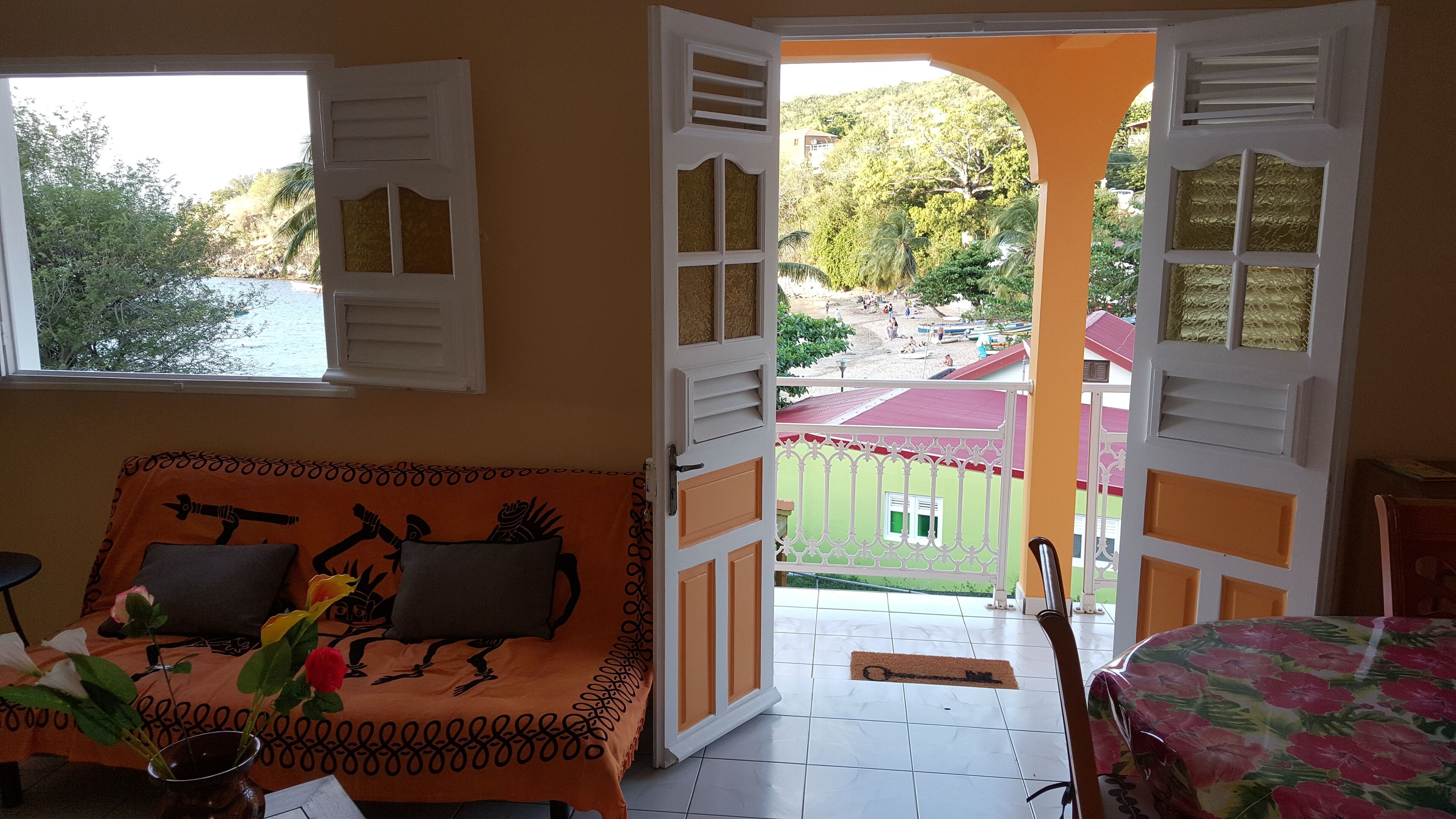 A room with a sofa, a small table, and a chair. There is a window with white shutters and a view of a green landscape with buildings.