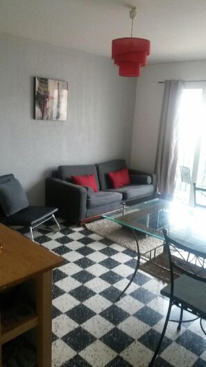 A living room with a checkered floor, a grey sofa with red cushions, a red pendant light, and a painting on the wall.