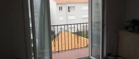Une pièce avec un sol à carreaux, une porte-fenêtre coulissante donnant sur un balcon doté d’une rambarde en métal, et un petit meuble placé à droite.