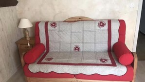 Ein Holzsofa mit roten Kissen und einer gesteppten Rückenlehne, platziert vor einer schlichten Wand. Daneben steht ein kleiner Nachttisch aus Holz.