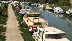 A canal lined with boats and RVs, a dirt path, and trees in the background. - Charming house completely renovated with swimming pool in the heart of the village for 4 people (Capestang)