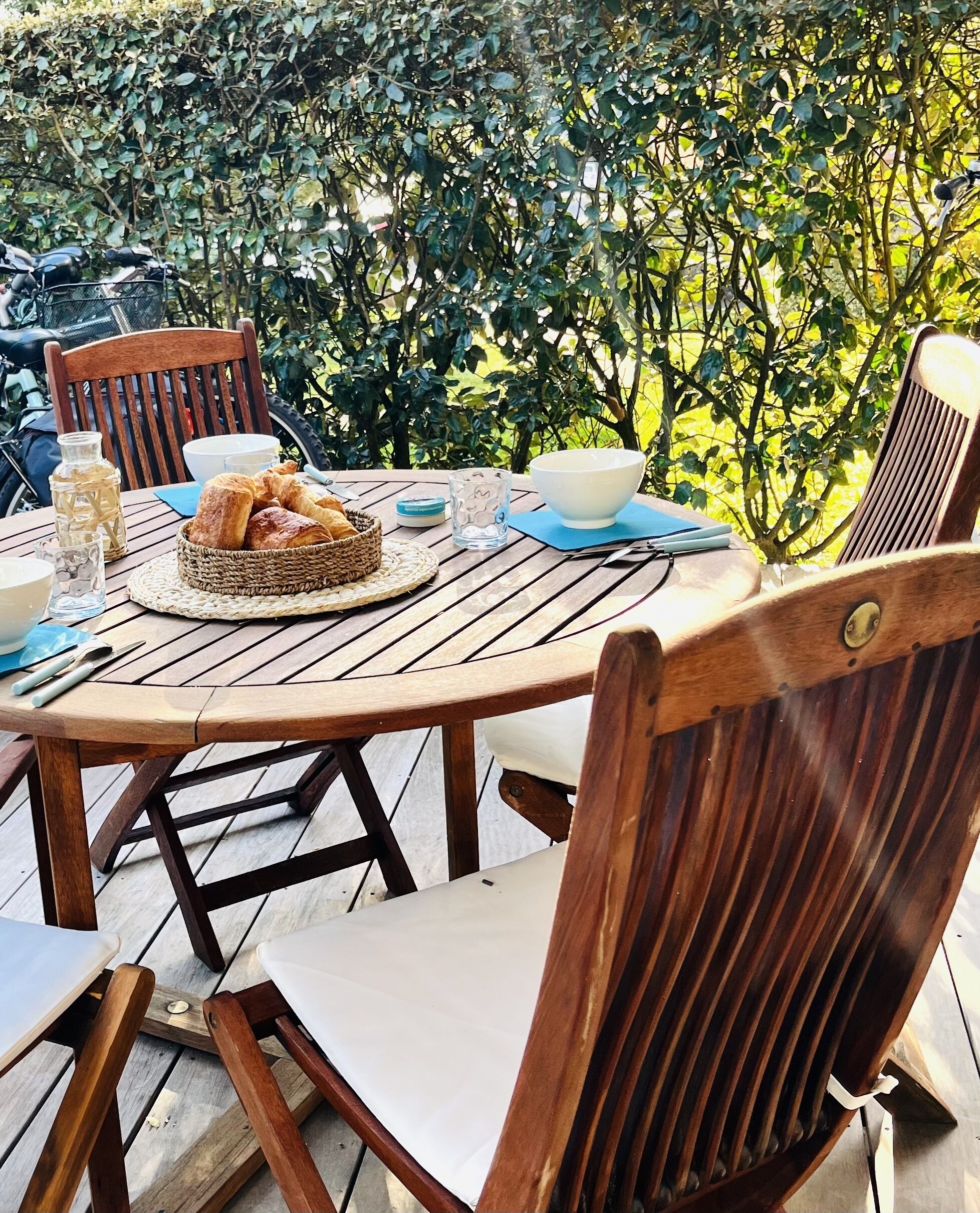 Una mesa redonda de madera al aire libre con sillas, una cesta de pan, un vaso de agua y un bol.