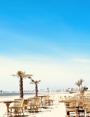 A beachside restaurant with wicker chairs and tables, palm trees, and a clear view of the ocean and city skyline in the background. - NEW in LABAULE! Near the beach, quality flat for 4 people,garden,swimming-pool (La Baule-Escoublac)