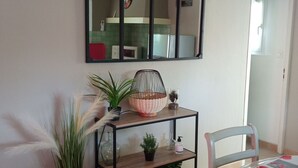 A room with a wooden floor, a metal shelf with plants, a mirror, and a chair with a floral tablecloth.