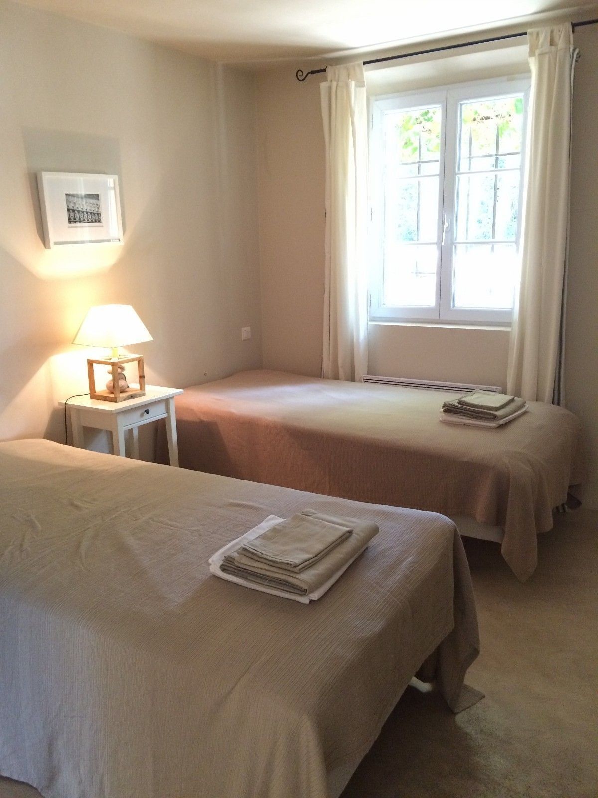 A room with two beds, a bedside table with a lamp, a window with curtains, and a framed picture on the wall.