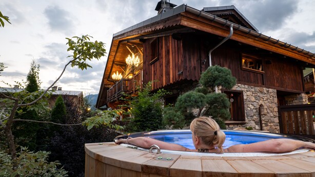 Een houten blokhut met een jacuzzi voor, omringd door bomen en een bewolkte lucht.