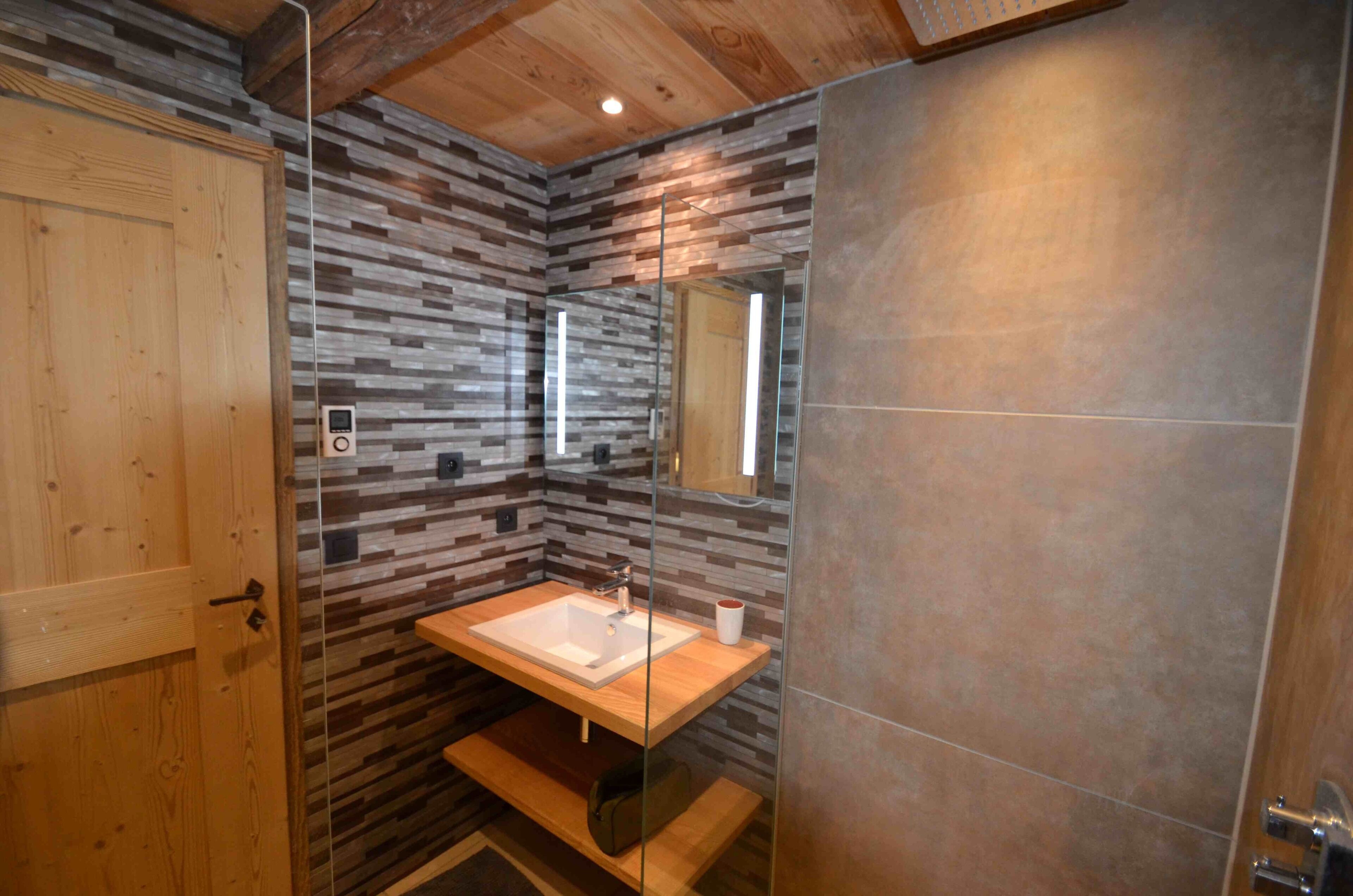 A modern bathroom with a wooden door, a glass-enclosed sink, and a tiled wall.