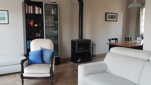 A living room with a white sofa, a chair with blue cushions, a bookshelf, a black stove, and a dining table with chairs. - House near paths and the Ria d'Etel 6 PERS. (Plouhinec)