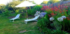 Un jardín con dos sillones blancos bajo una sombrilla blanca, rodeado de diversas plantas en flor.