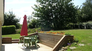 Terraza de madera con una mesa y sillas, rodeada por un jardín con flores y un paraguas rojo.