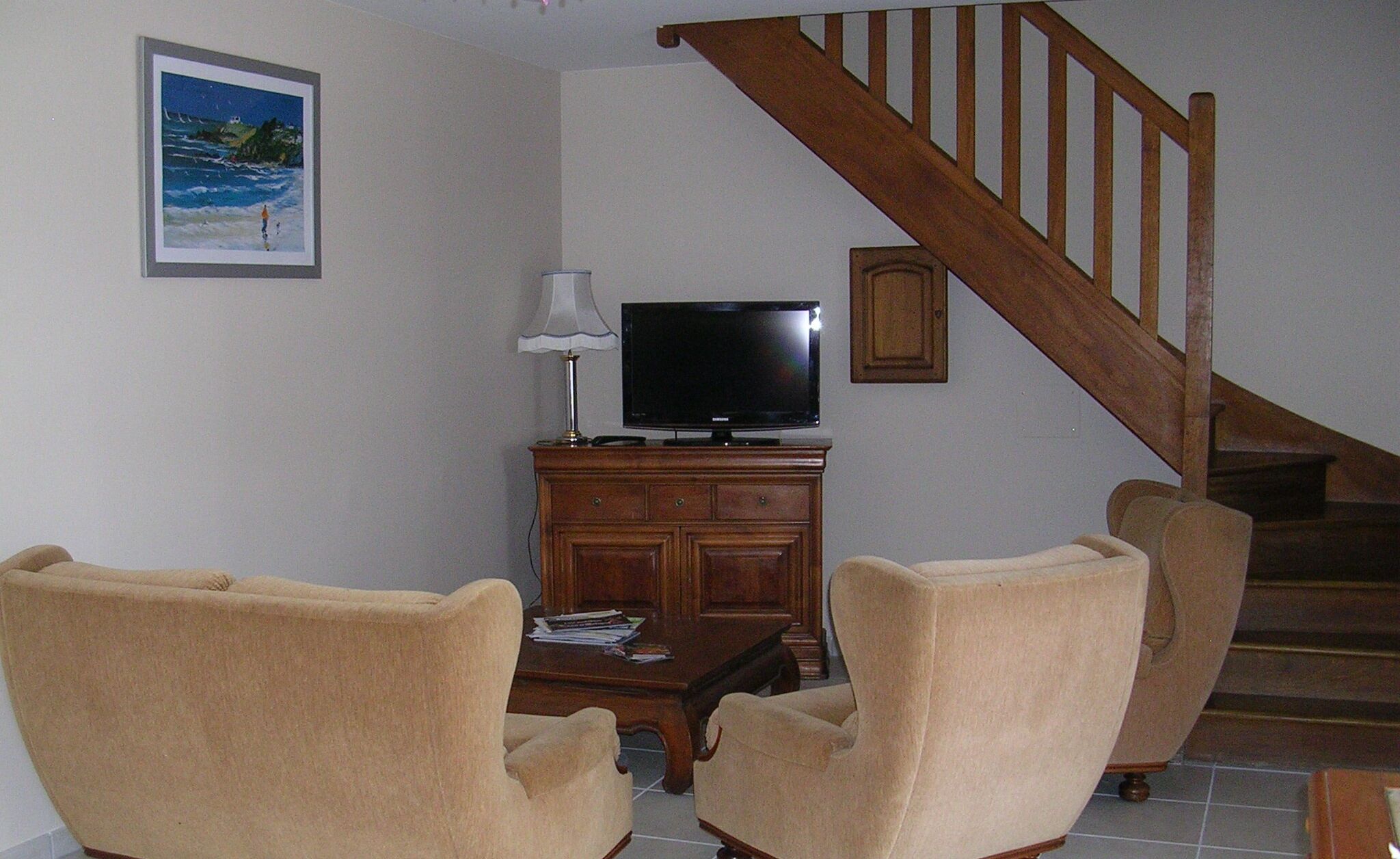 A living room with a television on a wooden cabinet, two beige armchairs, a staircase, and a framed picture on the wall.