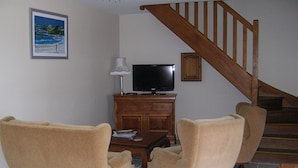 Una sala de estar con un televisor sobre un mueble de madera, dos sillones color beige, una escalera y un cuadro enmarcado en la pared.