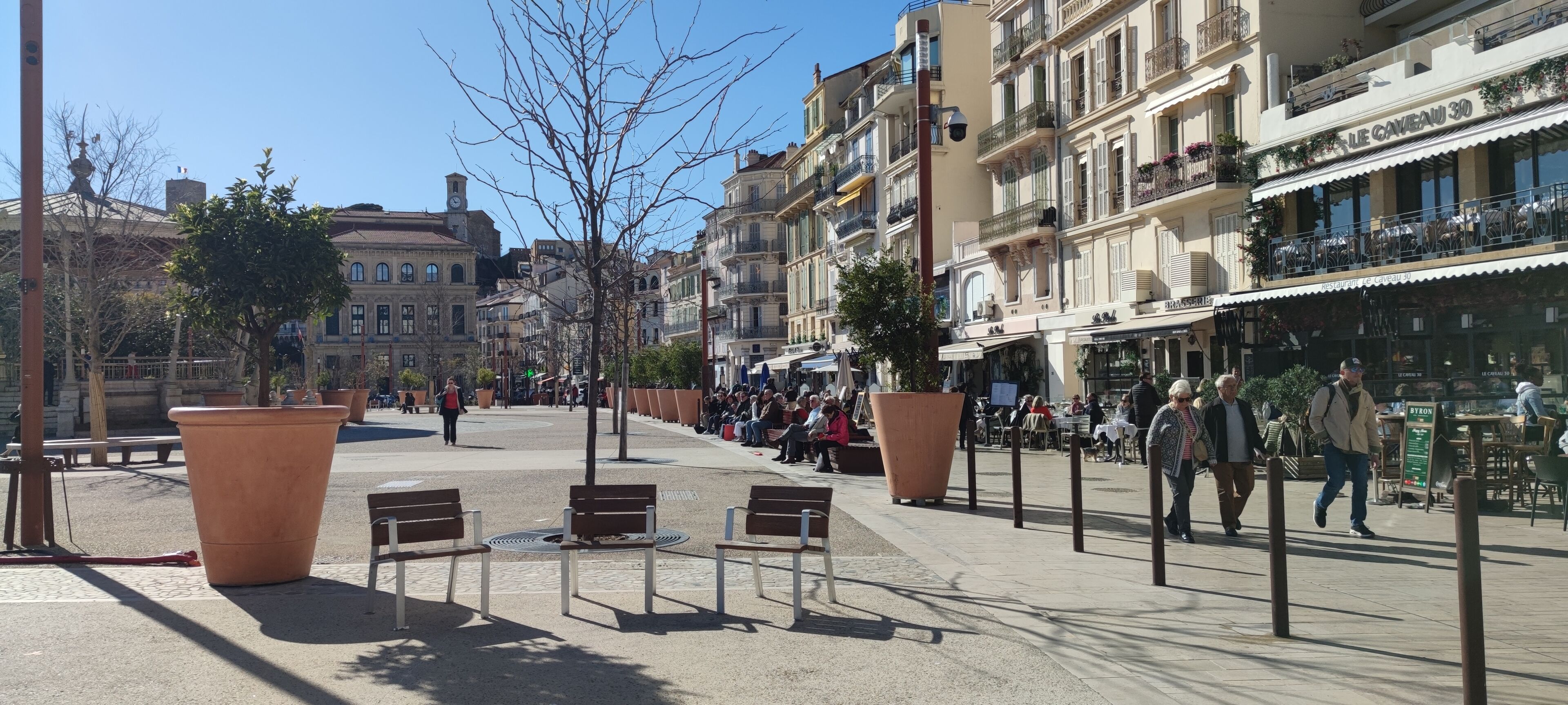A public square with benches, a clock tower, and buildings with balconies.