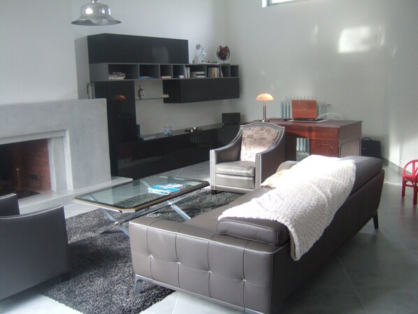 Una sala moderna con un sofá, un sillón y una mesa de centro de vidrio. Hay una chimenea, una estantería y un escritorio con una lámpara.
