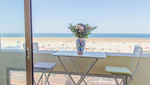 Un balcón con una mesa y dos sillas, con vistas a una playa con gente y sombrillas.