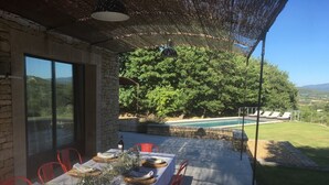An outdoor dining area with a table set for six, surrounded by red chairs, under a pergola with a view of a swimming pool and lush greenery.