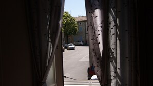 Et vindue med blomstrede gardiner, udsigt til en gade med parkerede biler, og en bygning i baggrunden.