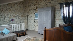 Et værelse med en stenvæg, en træseng, en hvid tremmeseng, en grå garderobe og et lille bord med en kuffert.