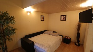 Ein Schlafzimmer mit einem Bett, einem Nachttisch, einem Sessel, einer Pflanze und einem an der Wand befestigten Fernseher.