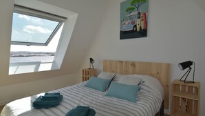 A bedroom with a skylight, a bed with striped bedding, two pillows, and a painting of a bus.